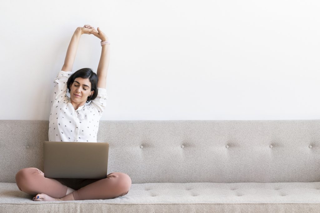 woman sitting on the couch with her laptop in her lap stretching her arms up