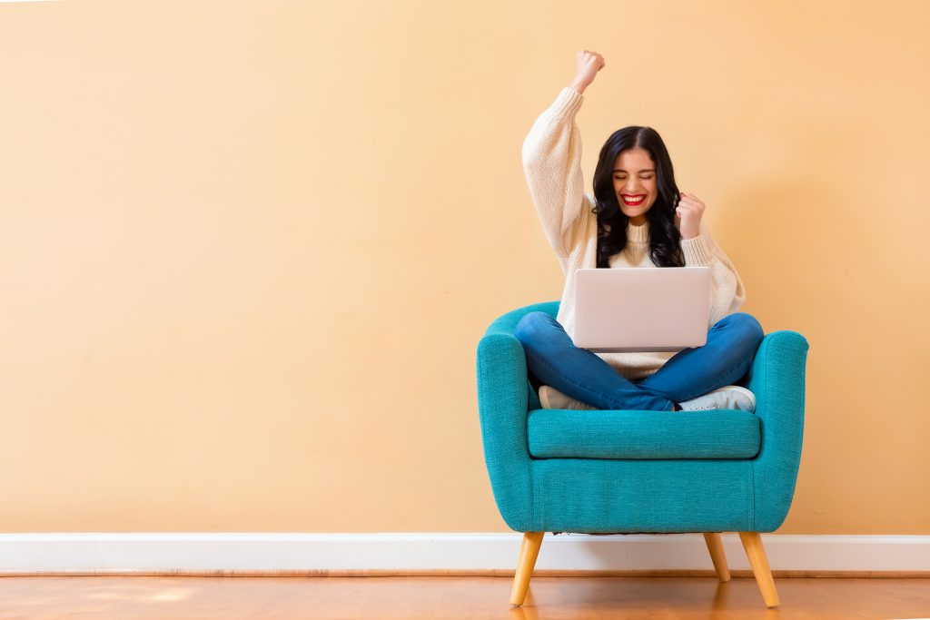Woman sitting on a blue chair with a laptop, raising her fist in success