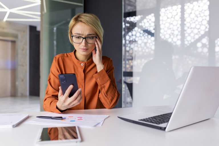 Sad upset woman pensive with phone in hands working inside office at workplace, businesswoman financier received notification message with bad news online, uses app on smartphone