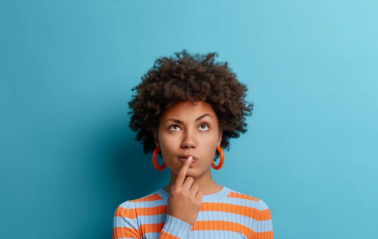 Vertical shot of thoughtful dark skinned woman has idea in mind, keeps finger on lips, looks pensively above, tries to decide how to act, poses against blue background, blank copy space above
