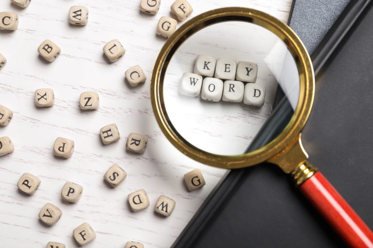 Looking at beads with word KEYWORD through magnifying glass at white wooden table, flat lay