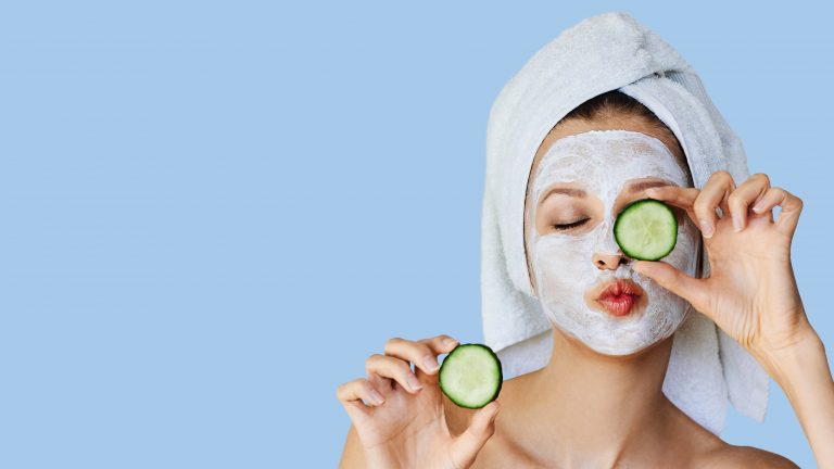 Young woman with facial mask on her face holding slices of fresh cucumber. Skin care and treatment, spa, natural beauty and cosmetology concept, over blue background with copy space
